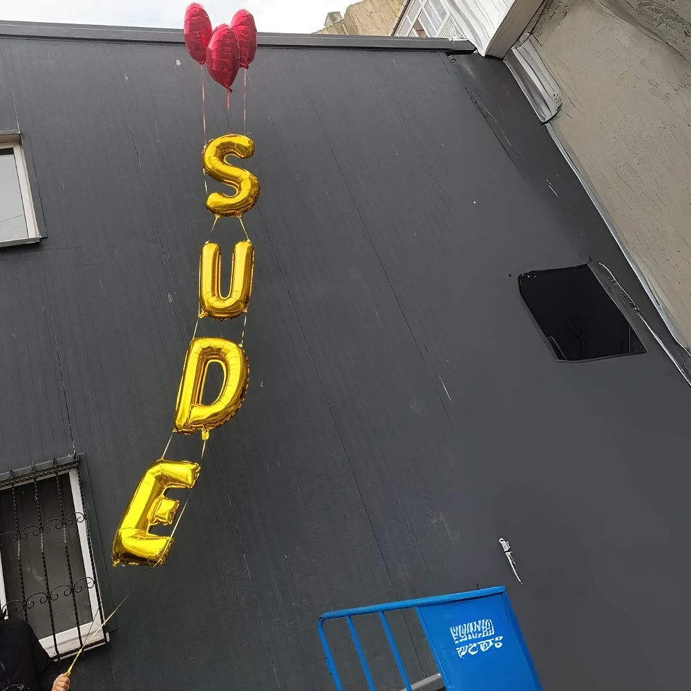 Altın harfli isimli uçan balon zinciri Altın renkli harf balonlarından oluşan dikey zincir, gri bir bina cephesinin önünde yükselirken alt ucundaki ip elde tutuluyor