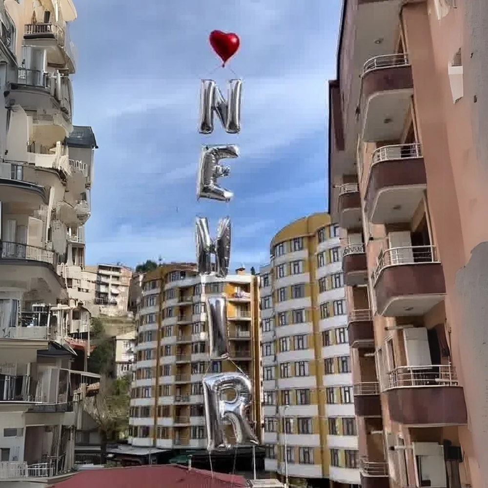 İsimli uçan balon seti: kalp balonun altında dikey harf zinciri Kırmızı kalp folyo balonun altına dikey olarak dizilmiş gümüş harf balonları, apartmanların arasında gökyüzüne yükseliyor