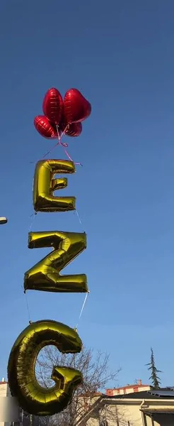 Gündüz çekilmiş altın harfli isim zinciri Mavi gökyüzüne yükselen altın renkli harf balonu zinciri, üstünde kırmızı kalp balonlar